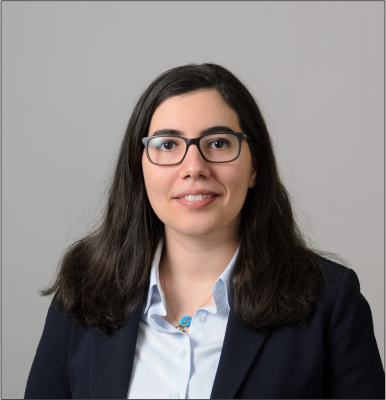 Professional headshot of a woman with dark hair and glasses, wearing a blazer and light blue shirt, against a neutral background.