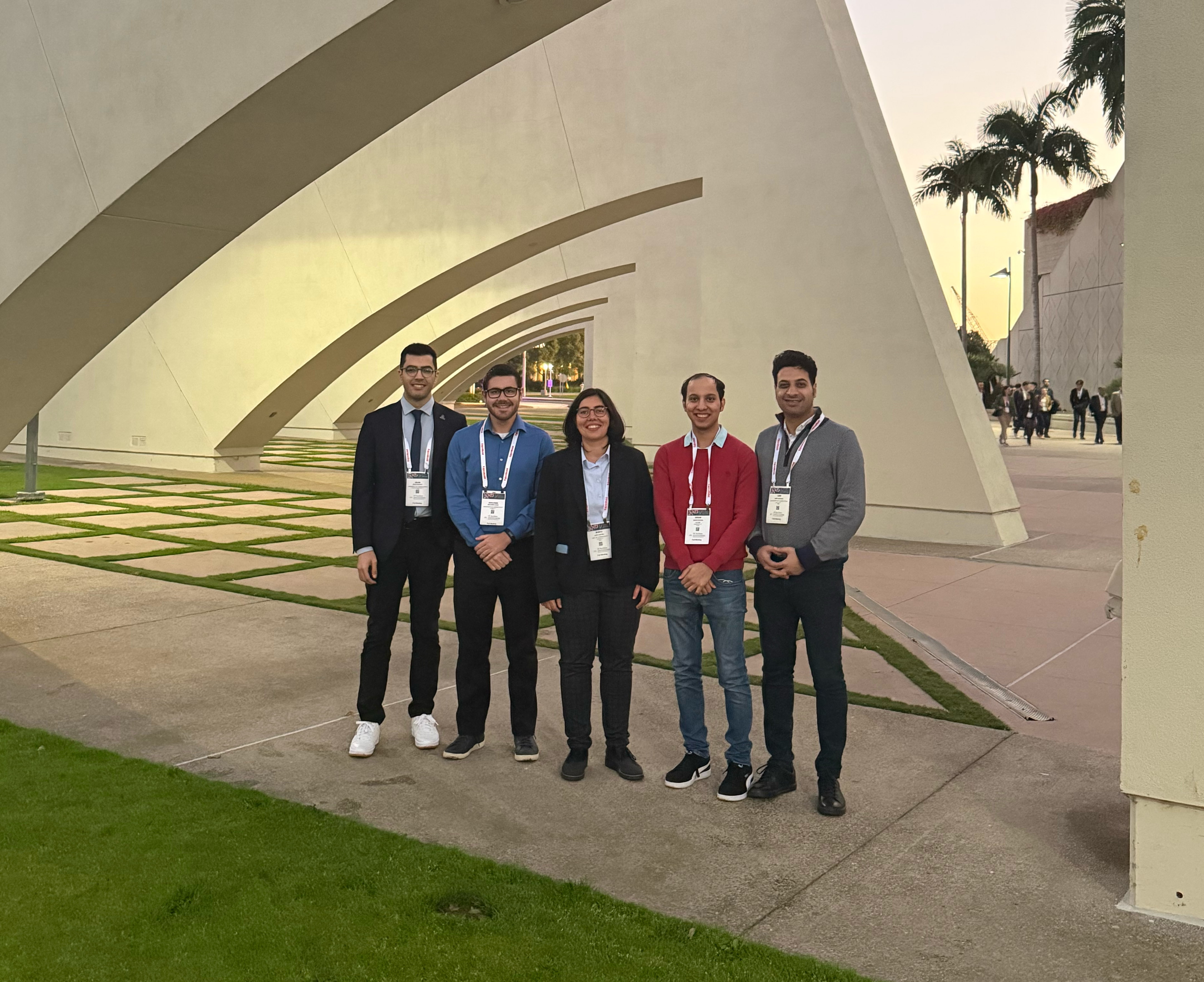 Group of five people posing outdoors near a modern arched building.